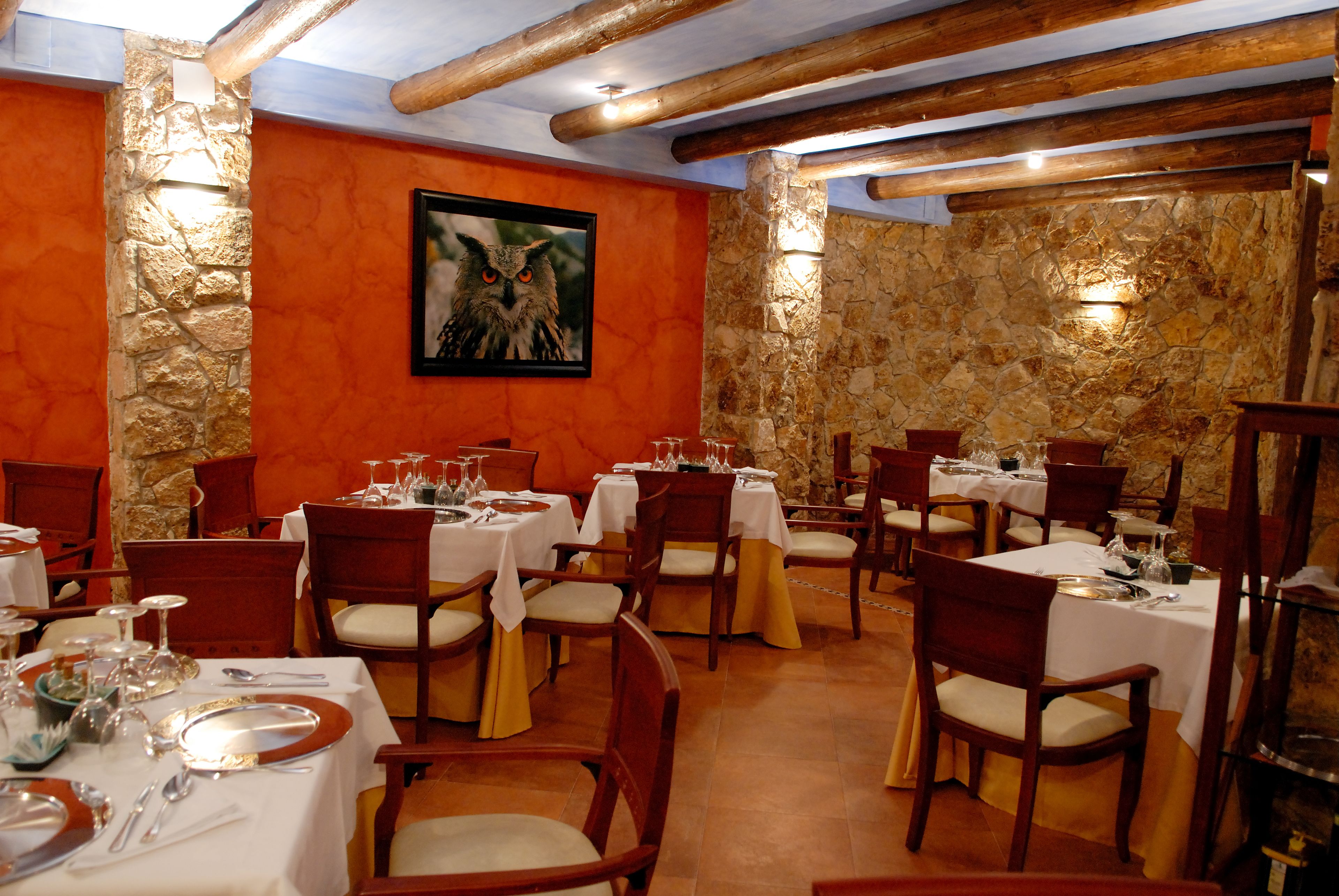 Un comedor con varias mesas dispuestas para una comida, paredes de piedra, vigas de madera y un cuadro enmarcado en la pared.