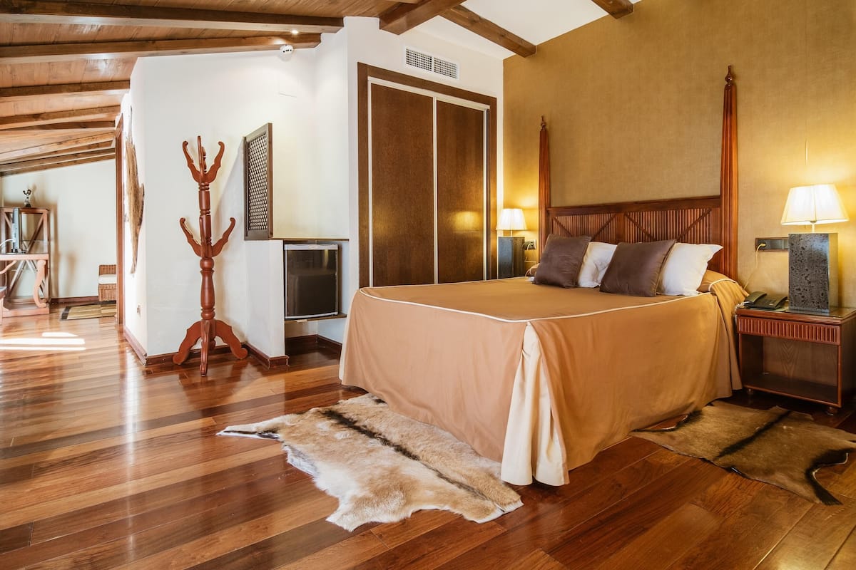 Un dormitorio espacioso con una cama grande, cabecero de madera y suelo de madera. Hay un perchero y un televisor en la esquina.