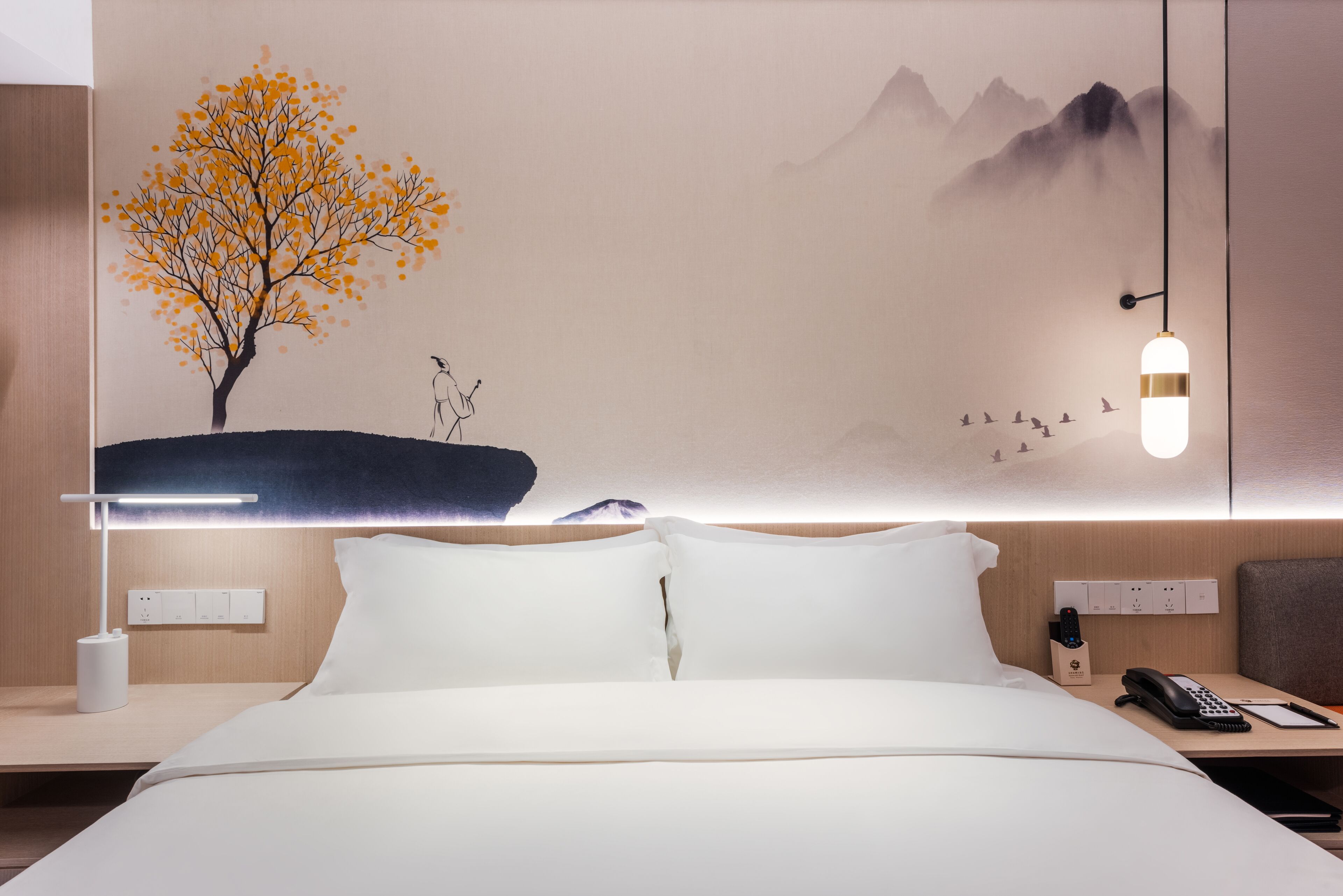 A hotel room with a bed, bedside table, lamp, and a wall mural of a tree and mountains.