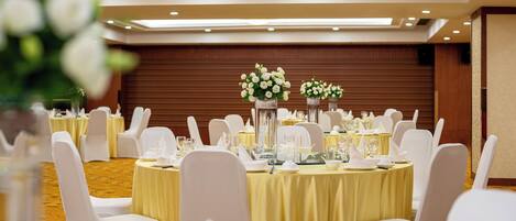 Un salón de banquetes con mesas redondas cubiertas con manteles amarillos, sillas blancas y centros de mesa florales.