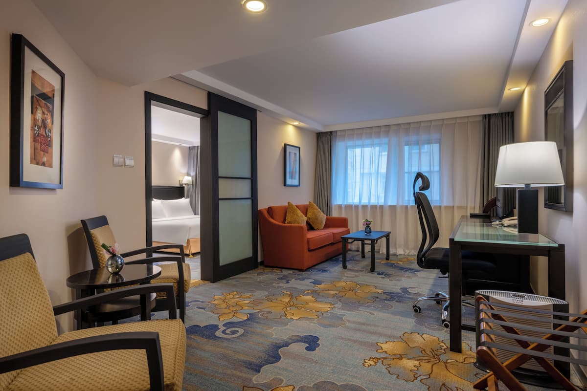 Une chambre d’hôtel avec un canapé, des fauteuils, un bureau, une lampe et une grande fenêtre avec des rideaux.