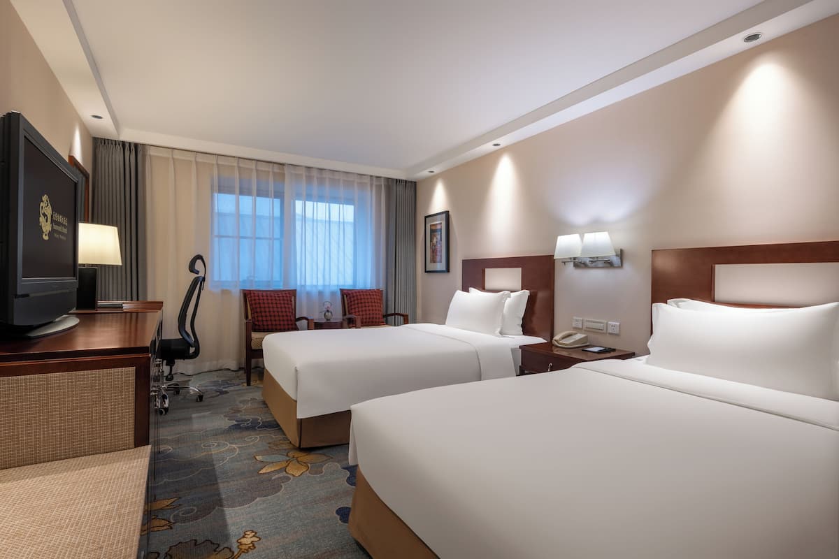 Une chambre d’hôtel avec deux lits, un bureau avec une télévision, une chaise, une lampe et une fenêtre avec des rideaux.