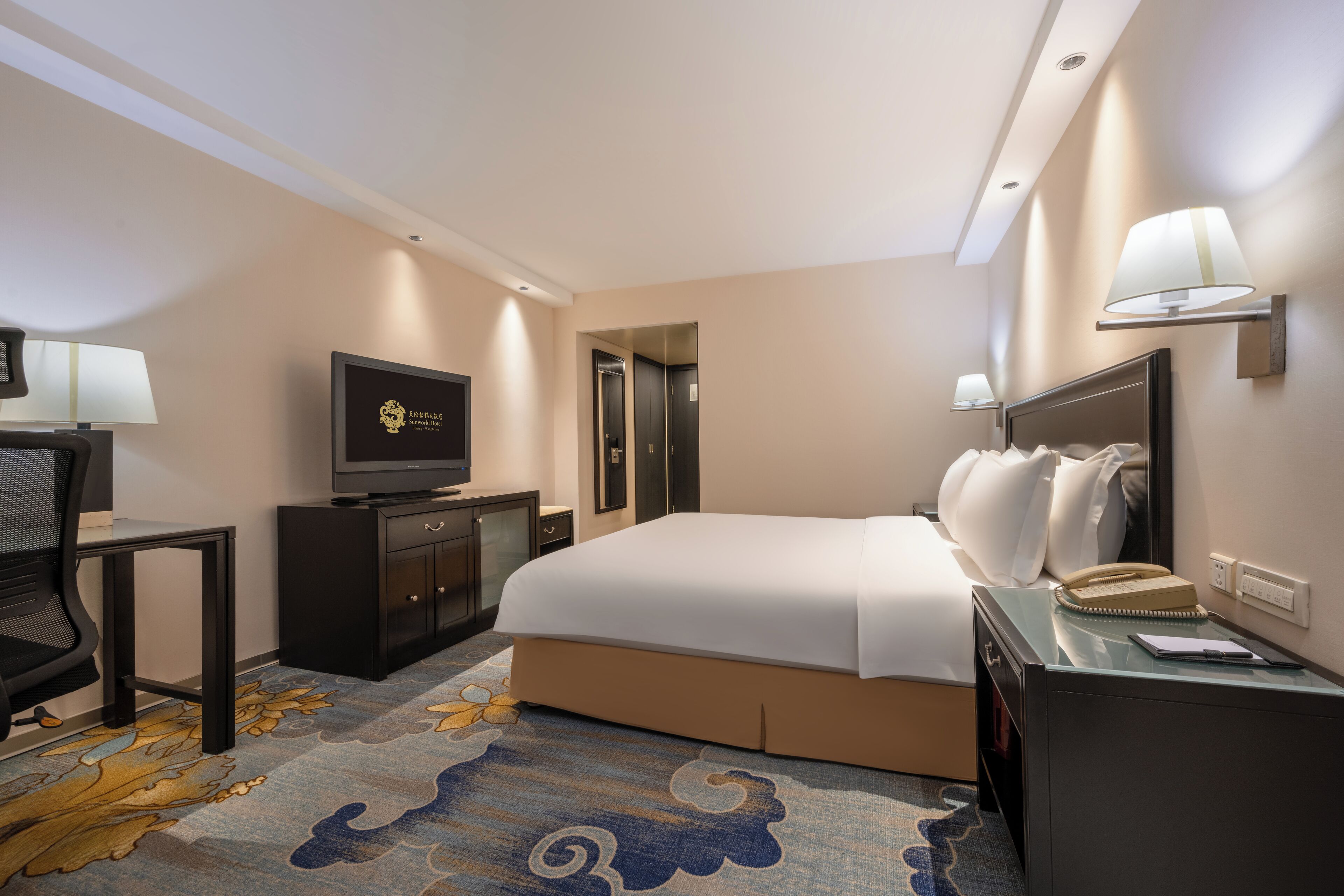 A hotel room with a bed, a desk with a chair, a television, a mirror, and a lamp.