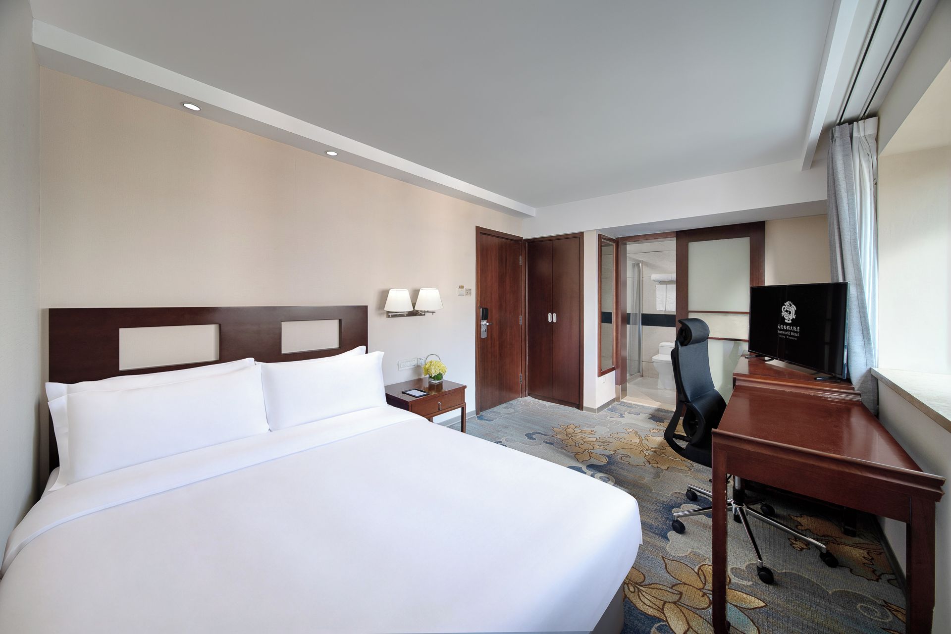 A hotel room with a large bed, a desk with a chair, a television, and a bathroom visible through an open door.