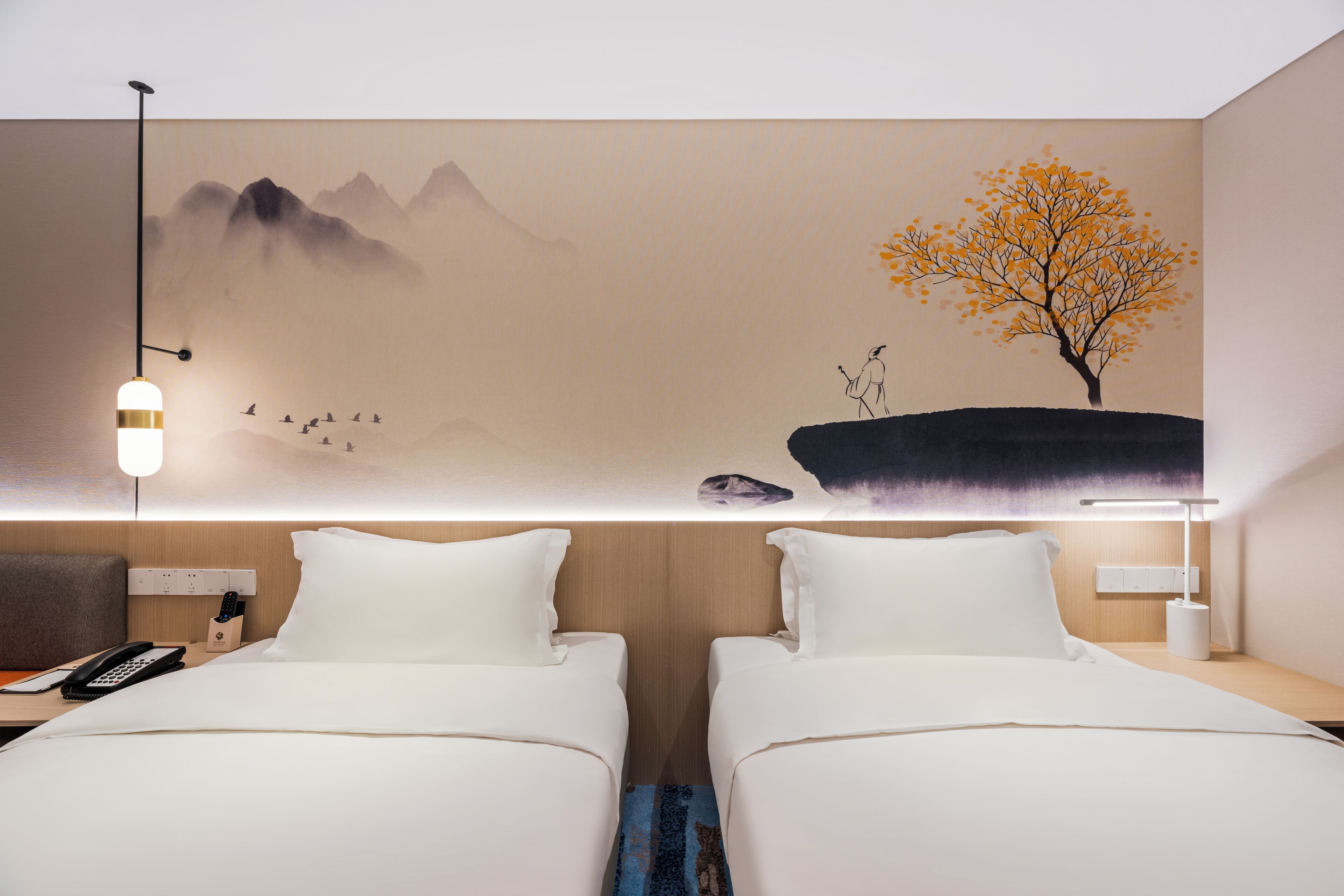 Une chambre d’hôtel avec deux lits, une table de chevet avec un téléphone, une lampe et une fresque murale représentant des montagnes et un arbre.