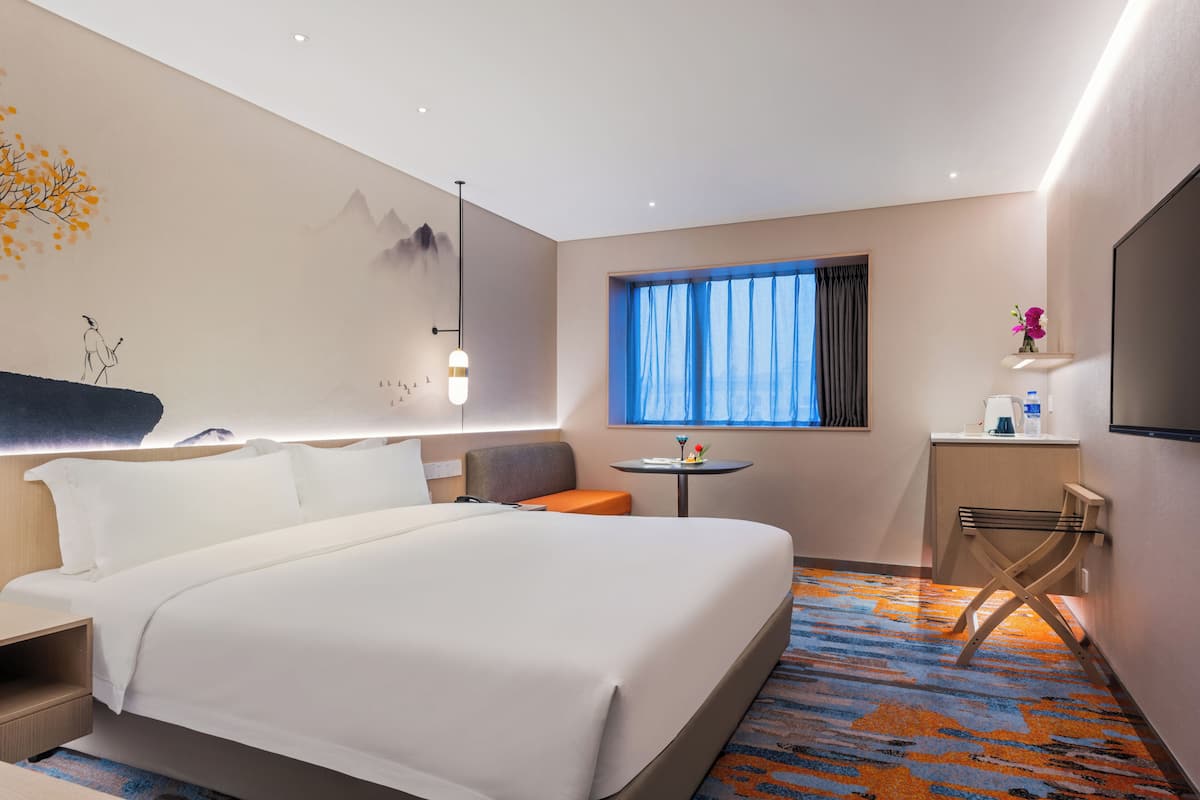 Une chambre d’hôtel avec un grand lit, un petit bureau, une chaise, une fenêtre avec des rideaux et un téléviseur fixé au mur.