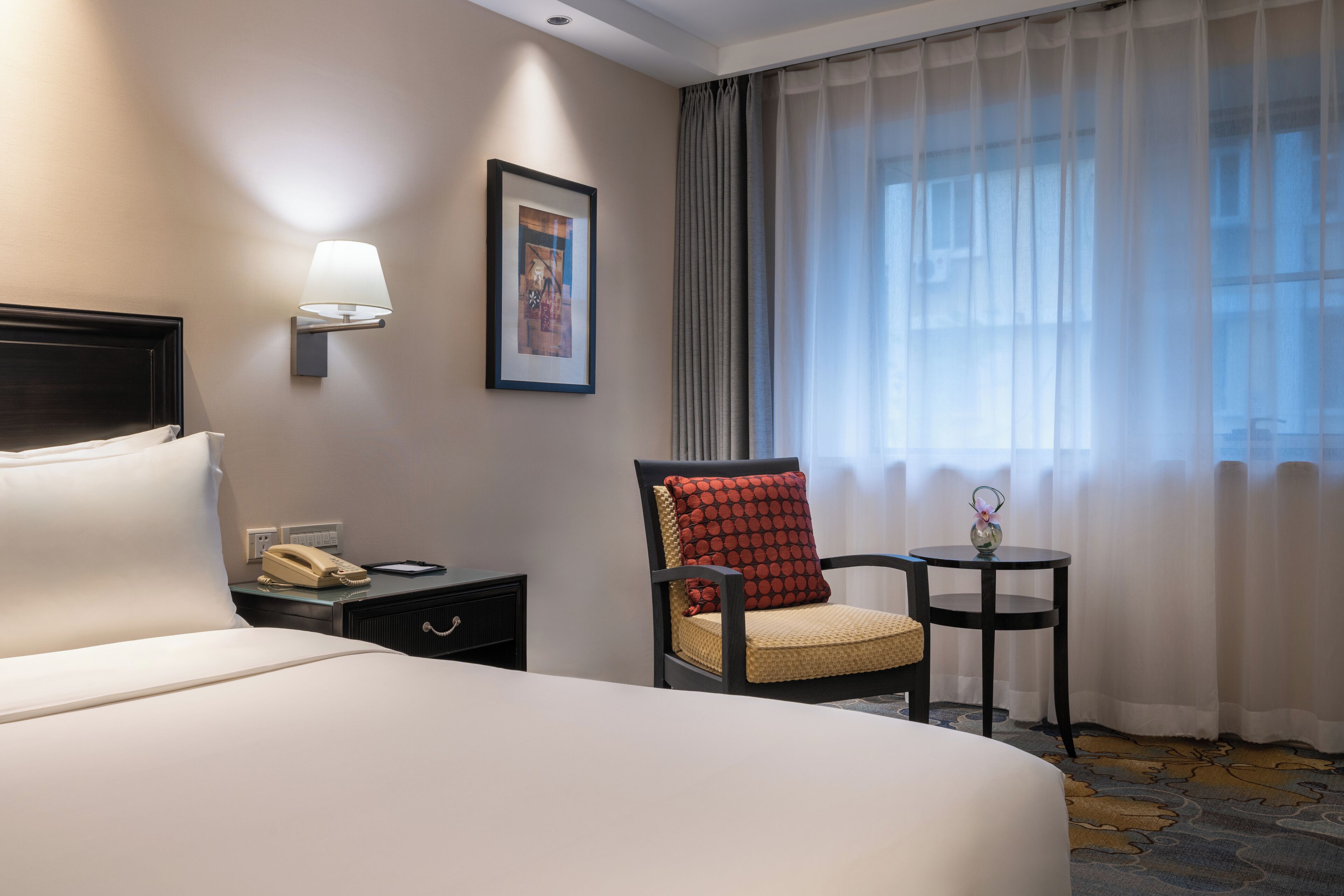 Een hotelkamer met een groot bed, een stoel met een rode kussen, een klein tafeltje met een vaas, een lamp en een ingelijst schilderij aan de muur.