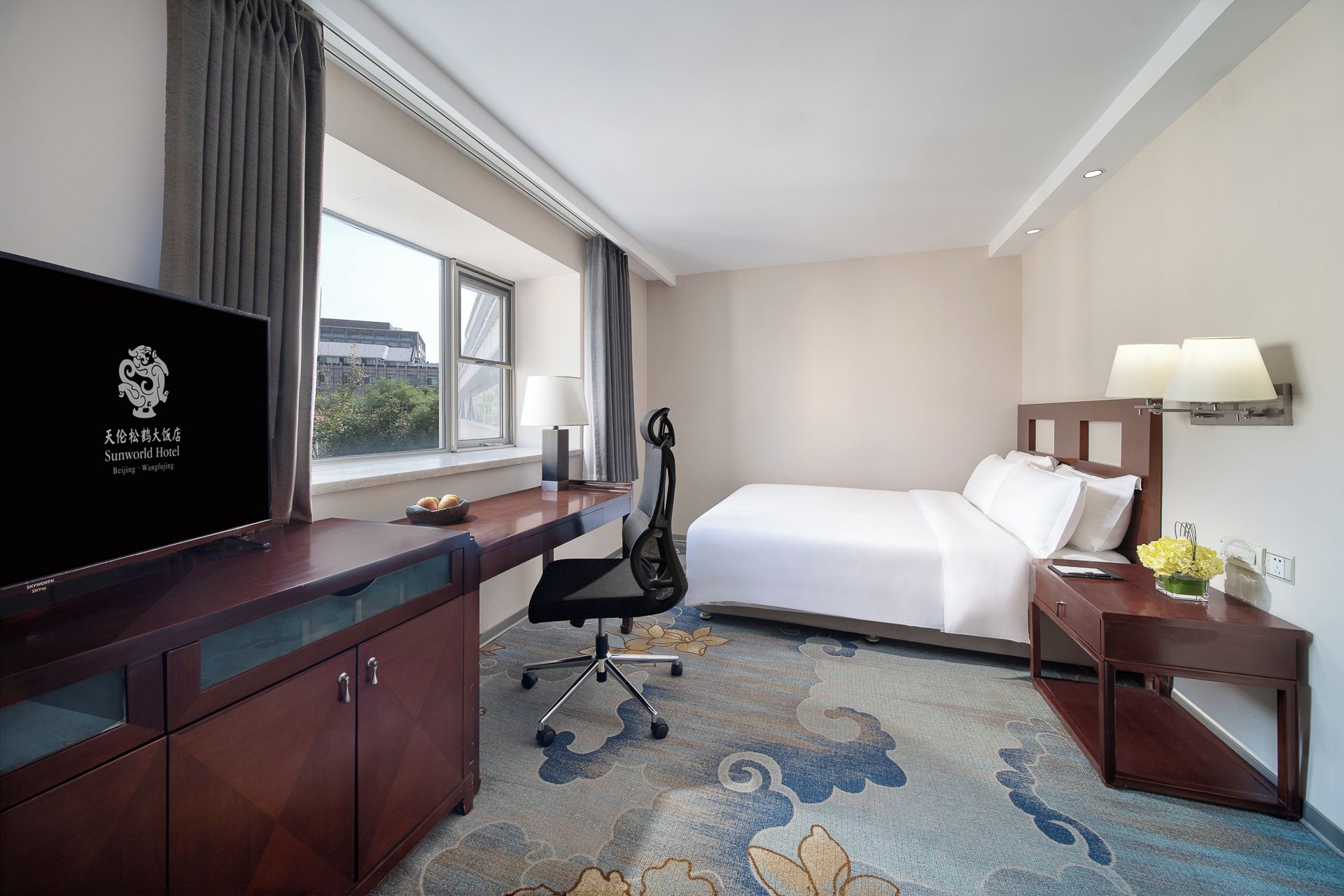 Une chambre d’hôtel comprenant un lit, un bureau avec une télévision, une chaise, une table de chevet avec une lampe et une fenêtre avec des rideaux.