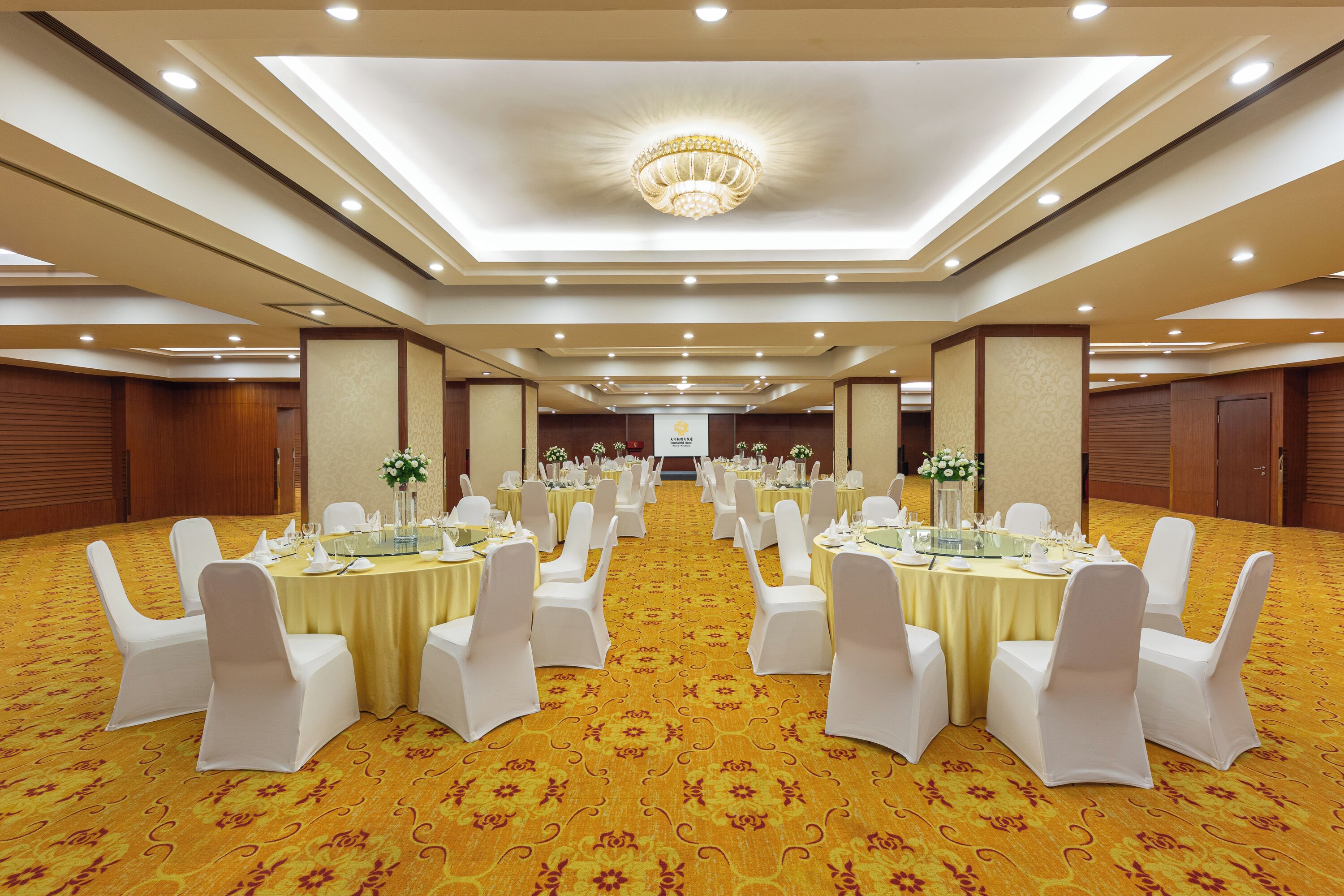 Un salón de banquetes con mesas redondas, sillas blancas y una alfombra amarilla.