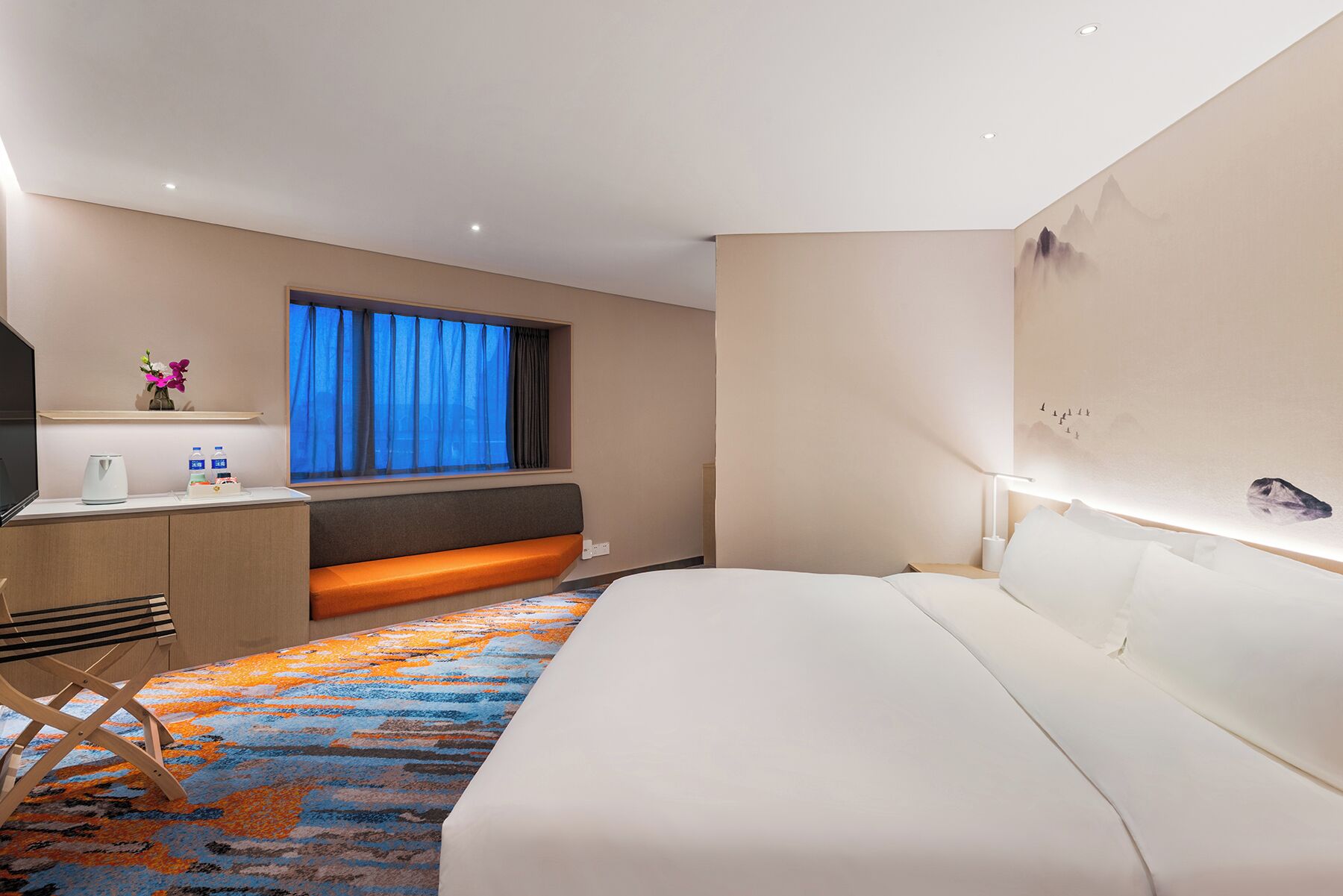 A hotel room with a bed, a desk with a chair, a window with curtains, and a wall with a decorative element.
