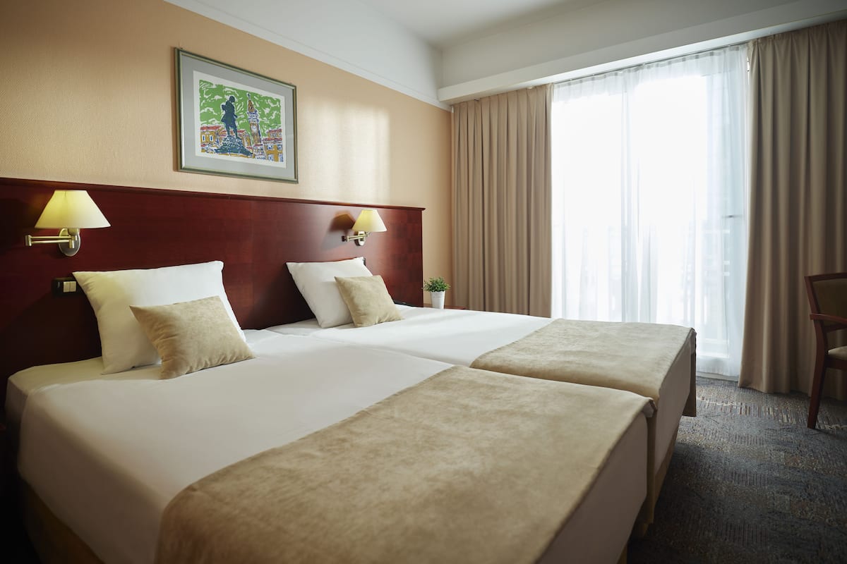 A hotel room with two beds, beige bedding, a wooden headboard, a framed cityscape picture, a window with curtains, and a small plant on the nightstand.