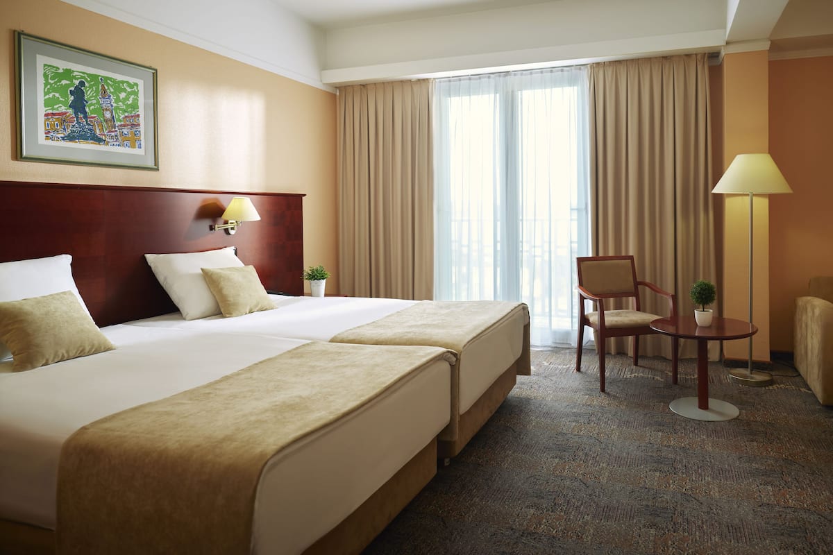 A hotel room with two beds, a desk, a chair, a lamp, and a painting on the wall.