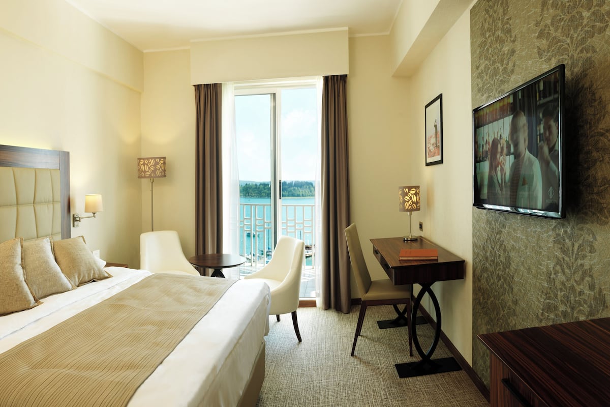 A hotel room with a large bed, a desk, a chair, and a TV. There is a balcony with a view of the water.