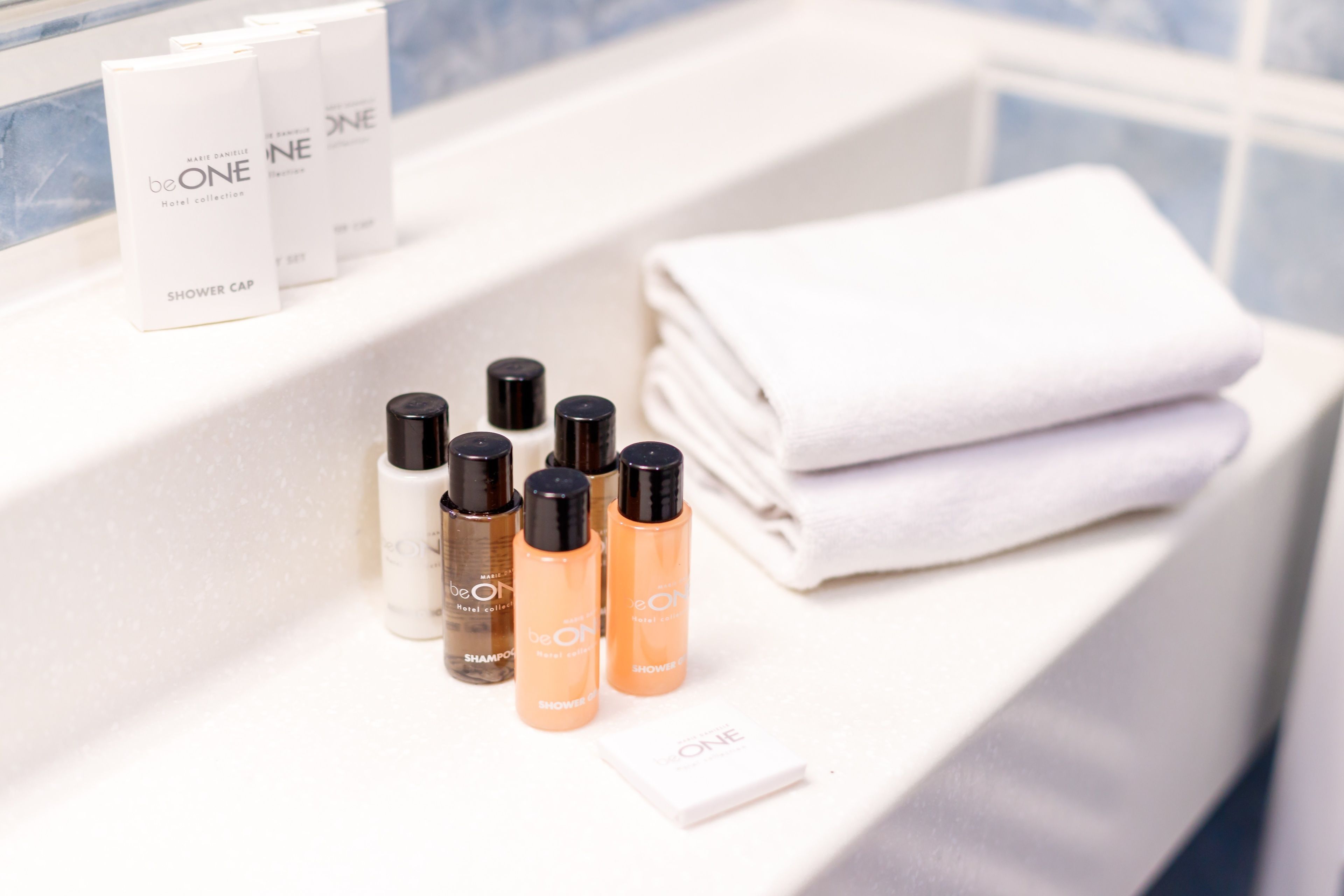 A bathroom counter with a stack of white towels, a folded white towel, and several bottles of Marie Daniele BeONE Hotel Collection products including shower caps, shampoo, and shower gel.