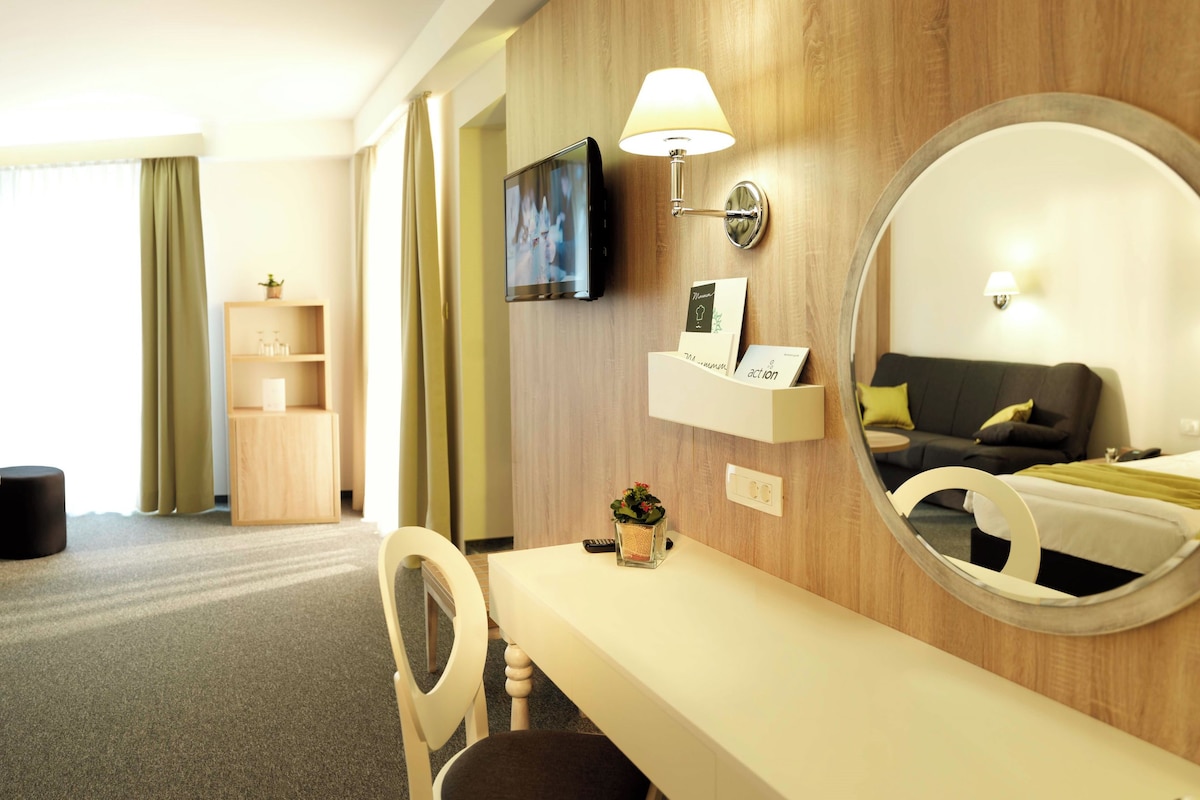 A hotel room with a bed, a sofa, a desk, a chair, a TV, a mirror, and a small table with a lamp.