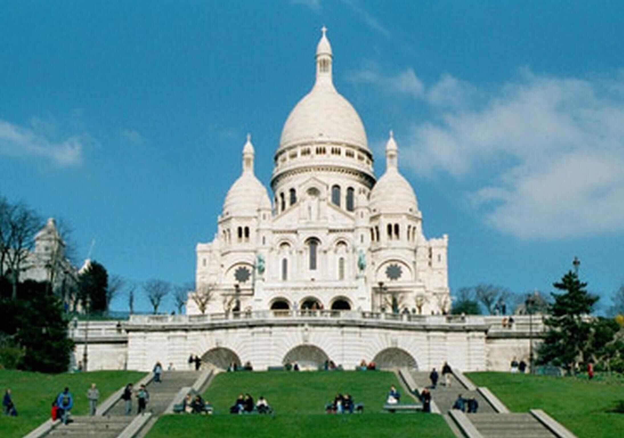 A large, white, domed building with multiple arches at the base, surrounded by a grassy area with people walking and sitting.