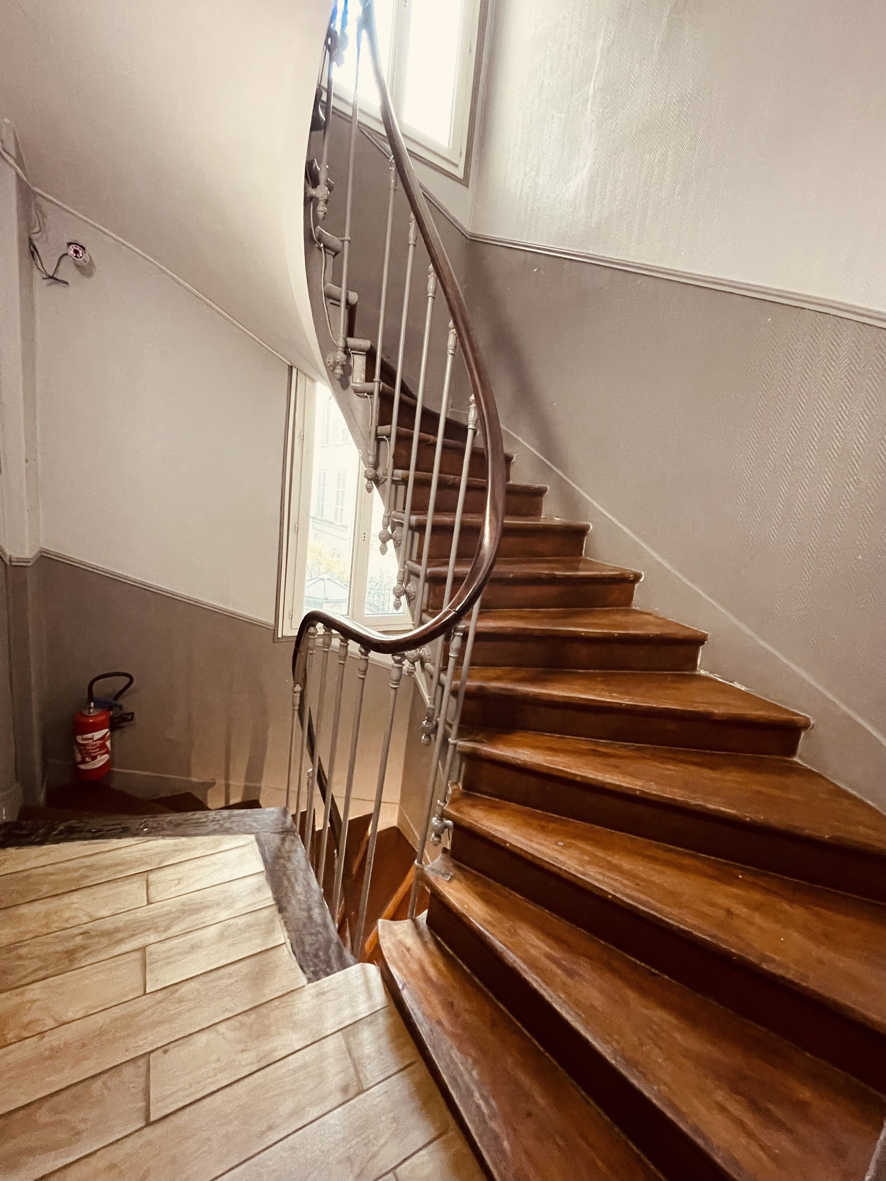Una escalera de madera con barandilla metálica, un extintor y una ventana en la plataforma.