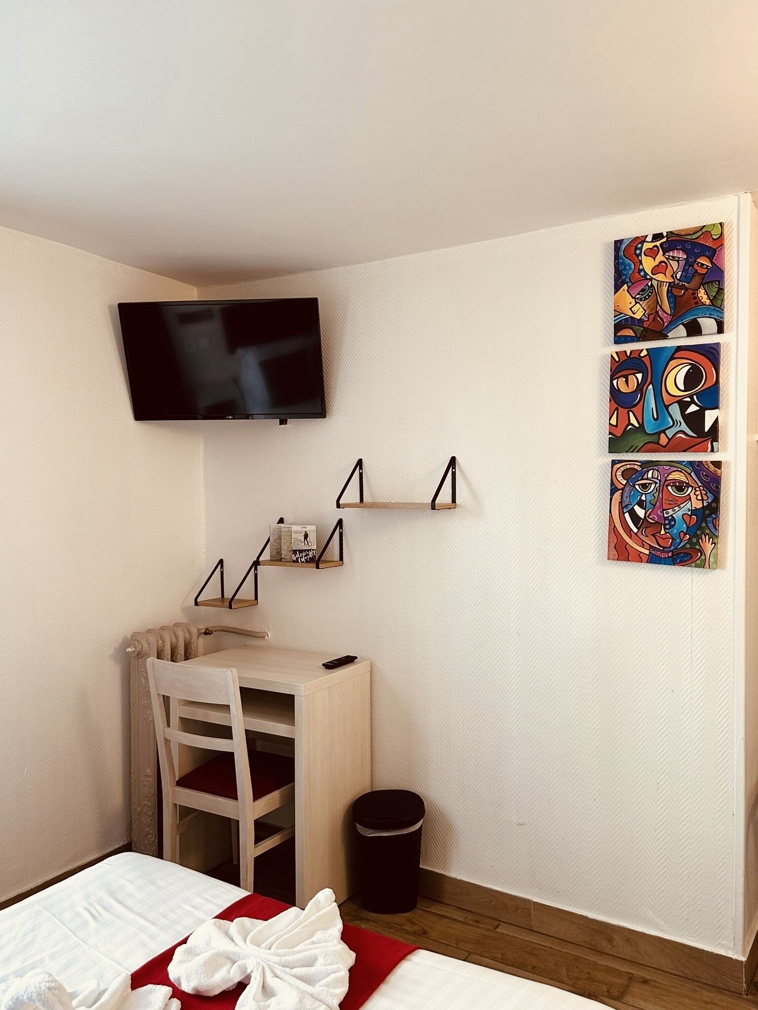 Quarto de hotel com uma cama, uma mesa com cadeira, televisão fixada na parede e obras de arte na parede.