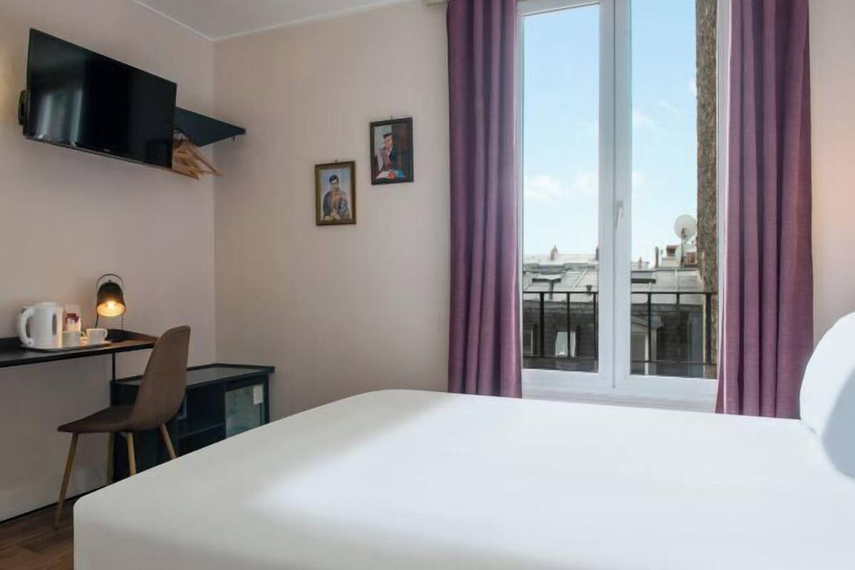 A hotel room with a bed, a desk with a lamp, a TV, and a view of the city.