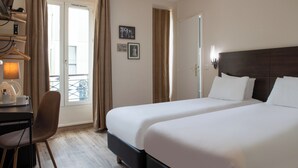 A hotel room with two beds, a desk with a chair, a lamp, and a view of the outside balcony. - Hôtel Istria by Magna Arbor (Paris)