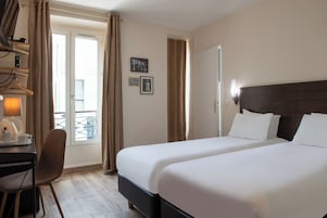 Habitación de hotel con dos camas, un escritorio con silla, una lámpara y vistas al balcón exterior.