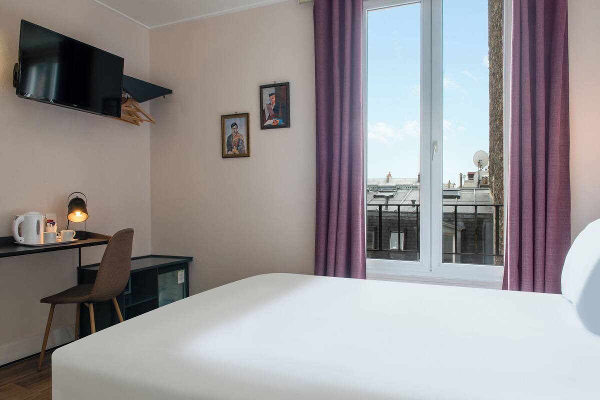 A hotel room with a bed, a desk with a lamp, a chair, and two framed pictures on the wall.