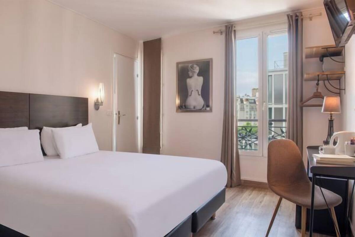 A hotel room with a bed, a desk with a chair, a painting on the wall, and a window with a view.