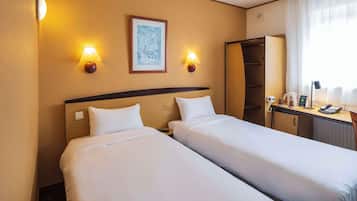 Two single beds with white linens and a wooden headboard, a desk with a telephone and lamp, and a wardrobe with a mirror.