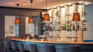 Una zona de bar con mostrador, taburetes y estantes llenos de botellas y vasos.