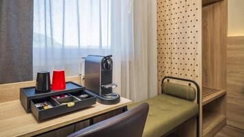 Habitación de hotel compacta con escritorio de madera, cafetera, un pequeño cajón con objetos, taza roja, banco acolchado verde y ventana con cortinas traslúcidas.