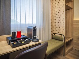 Habitación de hotel compacta con escritorio de madera, cafetera, un pequeño cajón con objetos, taza roja, banco acolchado verde y ventana con cortinas traslúcidas.