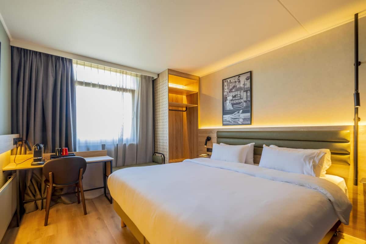 Quarto de hotel com uma cama grande, uma mesa e uma cadeira. Tem uma janela com cortinas, uma luminária e um quadro na parede.