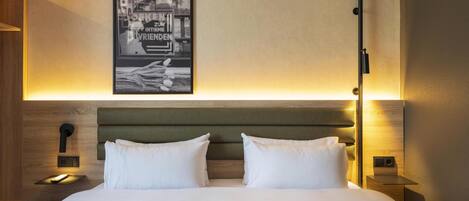 Habitación de hotel con una cama grande, dos almohadas, cabecero, mesitas de noche, una lámpara de pared y un cuadro enmarcado.