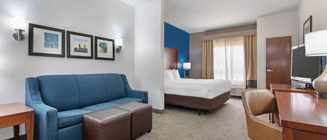 Une chambre d’hôtel avec un canapé bleu, un lit, un bureau et une chaise. Des tableaux encadrés ornent le mur, et il y a une fenêtre avec des rideaux.