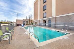 A swimming pool with a metal railing, green and white lounge chairs, and a building with a brick facade in the background.