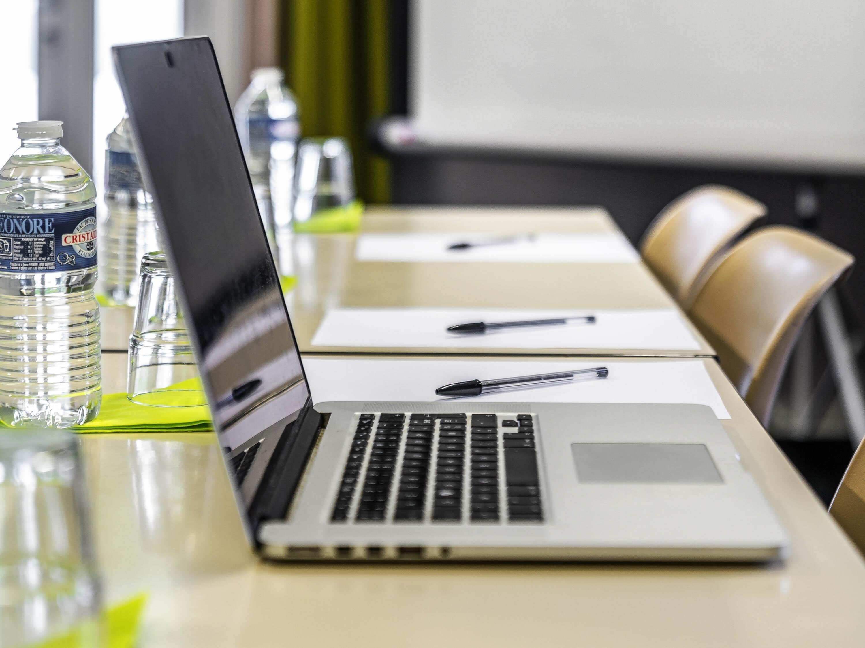 A laptop on a table with a bottle of water, a pen, and a projector screen in the background.
