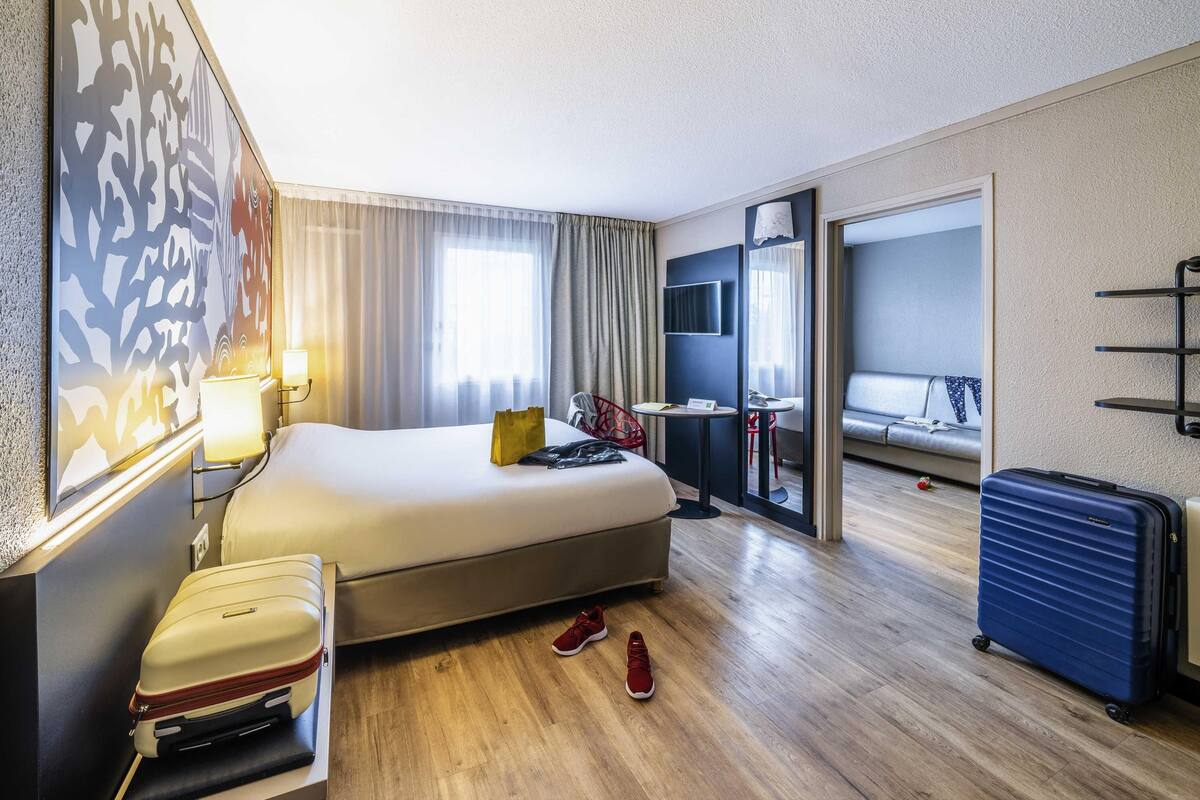 Una habitación de hotel con cama, escritorio, silla, televisor, ventana, puerta y maleta.