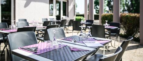 An outdoor dining area with tables and chairs, a building with large windows, and greenery in the background.