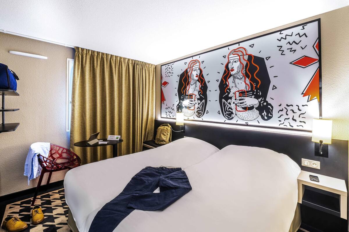 Habitación de hotel con cama, escritorio con computadora portátil, silla y un cuadro abstracto grande en la pared.