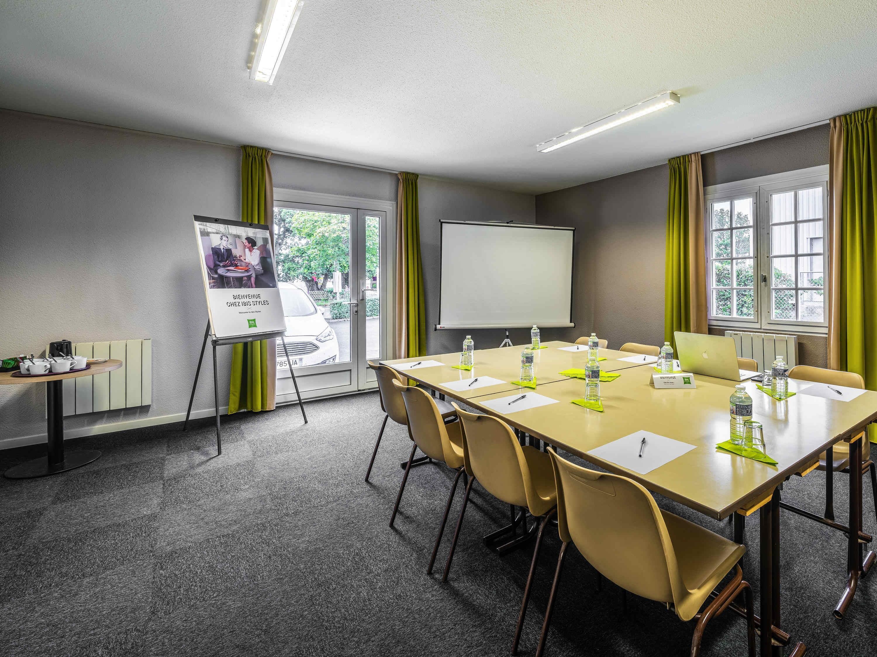 Une salle de conférence avec une longue table, des chaises, un écran de projection, un tableau blanc, une petite table avec des tasses et une bouilloire, et une affiche accrochée au mur.