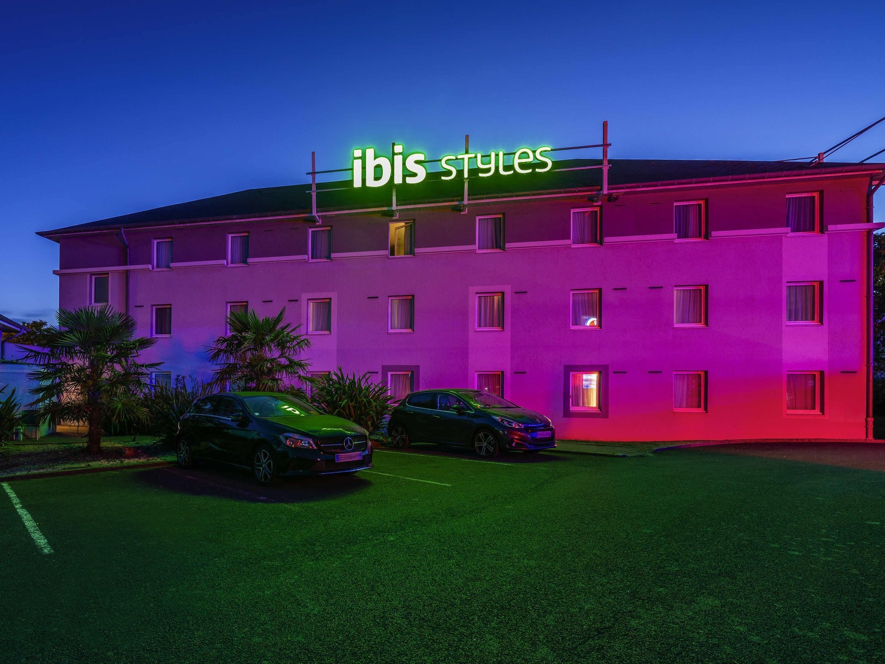 Et hotel med flere etager og et neonskilt med teksten 'ibis styles' på taget.