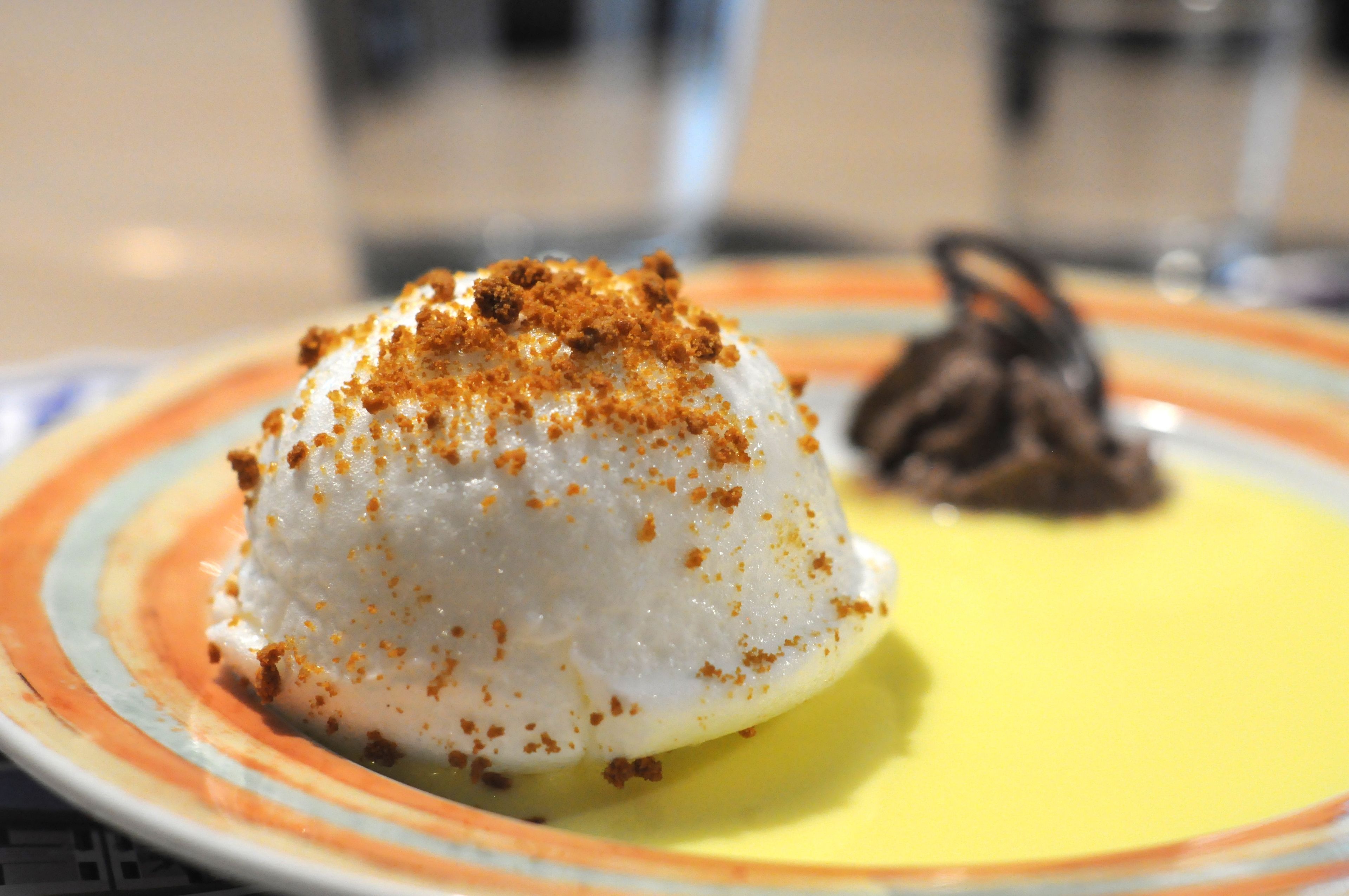 A dessert on a plate with a yellow base and a white mound topped with brown crumbs.