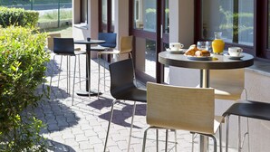 An outdoor seating area with tables and chairs, a clear view of the street, and a window with a view of greenery. - Comfort Hotel Linas Montlhery (Linas)