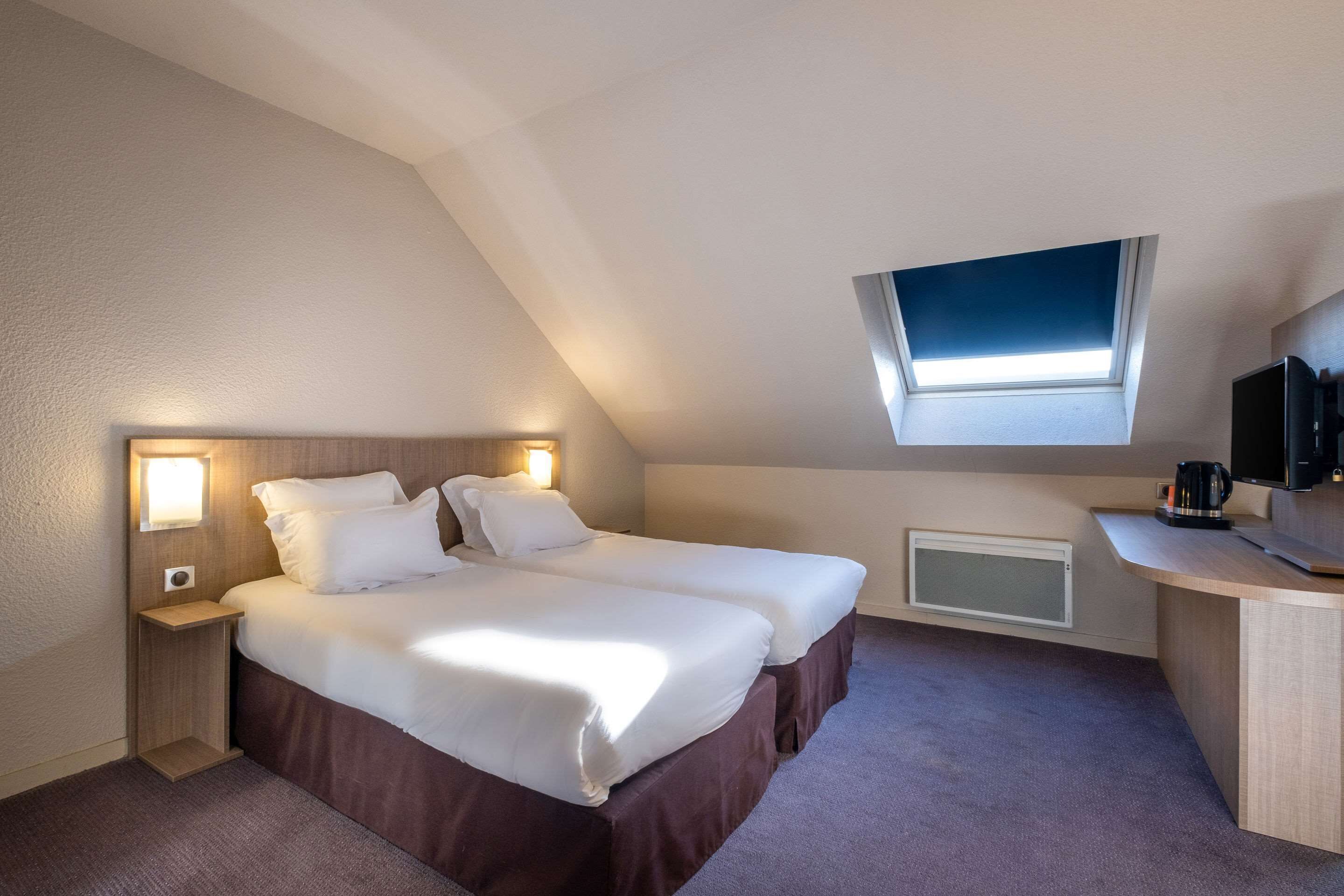 A hotel room with two beds, a desk, a coffee maker, a TV, and a skylight.