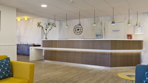 A reception area with a wooden reception desk, a clock, and a wall-mounted menu board. - Comfort Hotel Linas Montlhery (Linas)