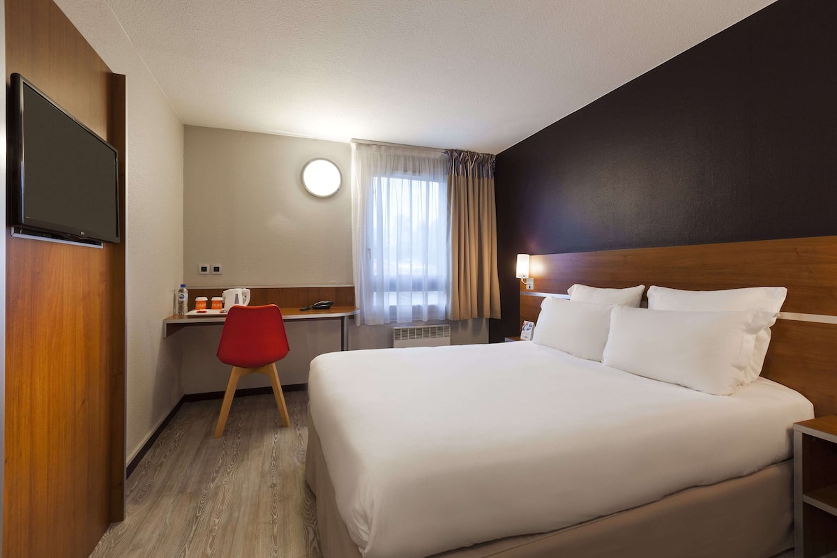 Una habitación de hotel con una cama grande, un escritorio con silla, un televisor y una ventana con cortinas.