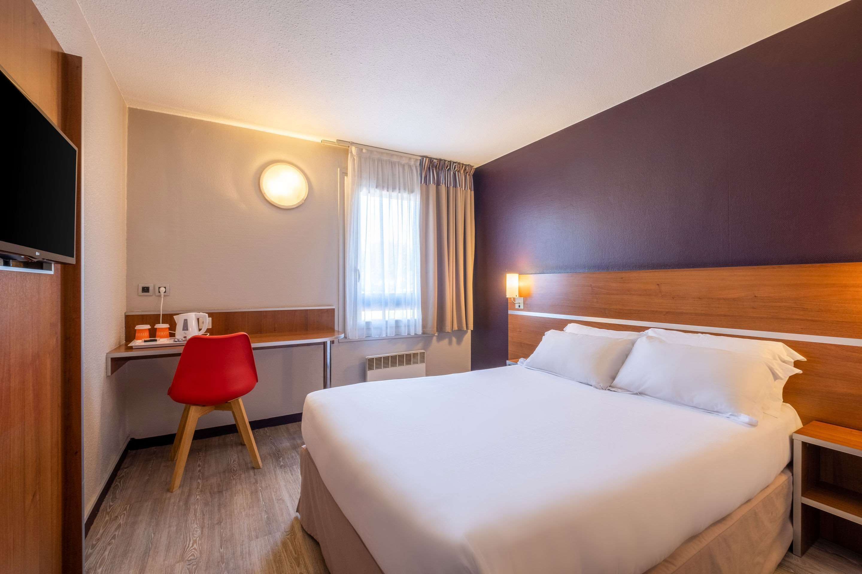Habitación de hotel con una cama grande, un escritorio con una silla roja, televisión y ventana con cortinas.