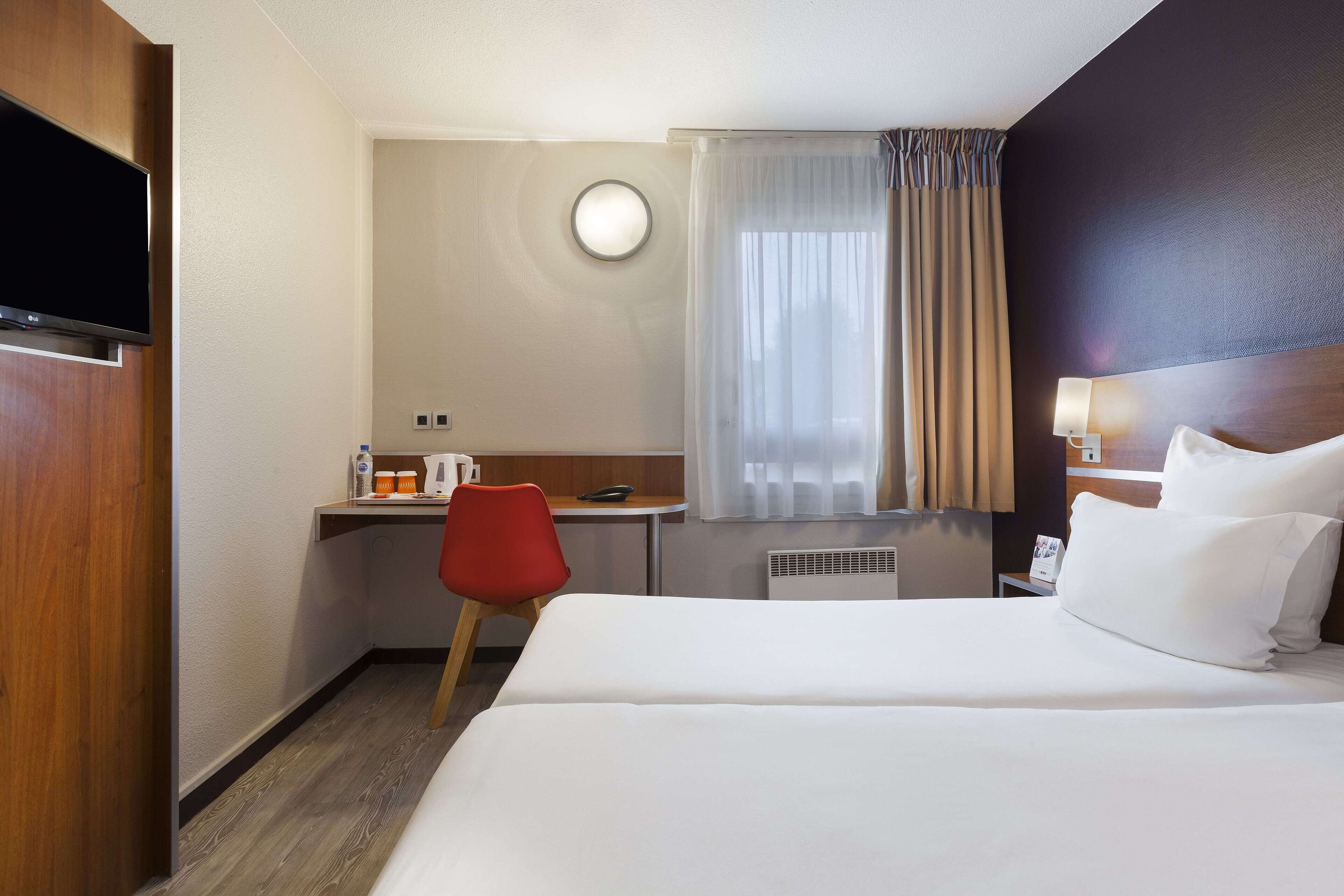 Ein Hotelzimmer mit Bett, Schreibtisch, Stuhl, Fernseher, einem Fenster mit Vorhängen und einer Wandlampe.