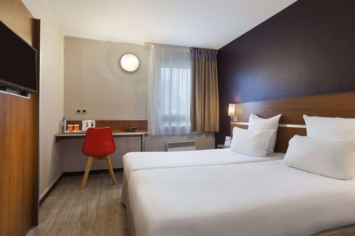 Una habitación de hotel con dos camas, un escritorio con silla, un televisor y una ventana con cortinas.