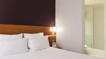 Een net opgemaakt bed met witte lakens, een houten hoofdbord en een nachtkastje met een lamp en een brochure.